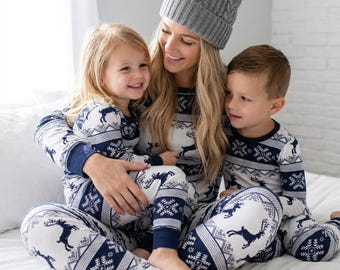 May include: Matching white and navy blue long-sleeve pajama sets featuring a repeating pattern of reindeer and snowflakes. The set includes a gray knit beanie with a pom-pom on top. The pajamas have a crew neckline and fitted cuffs at the wrists.