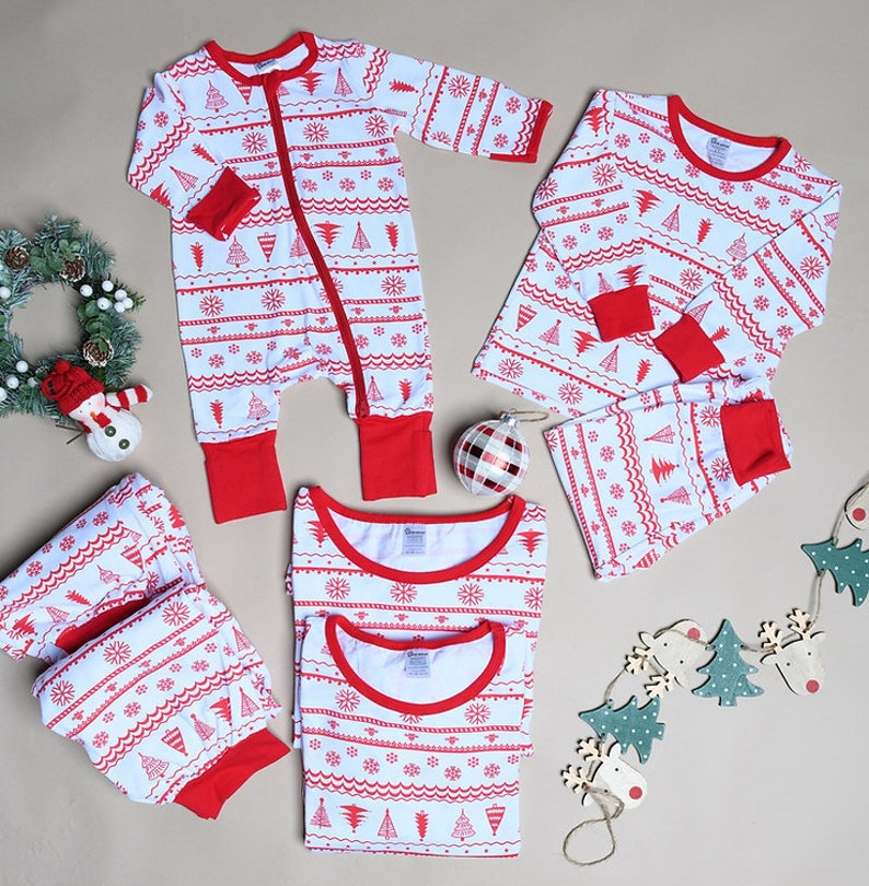 May include: A collection of white pajamas featuring a red pattern of Christmas trees, snowflakes, and wavy lines. The set includes a long-sleeve romper with a zipper, two-piece pajama sets with long sleeves and pants, all with red cuffs and necklines.