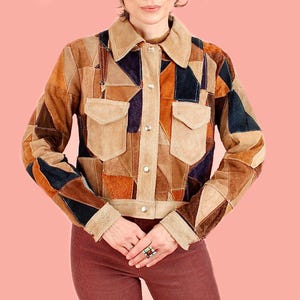 May include: A cropped jacket constructed from a patchwork of suede and leather pieces in various shades of brown, tan, and black. The jacket features a pointed collar, long sleeves with buttoned cuffs, and two chest pockets with flaps and silver buttons.