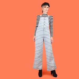 May include: A light blue denim overall featuring diagonal stitching patterns across the legs and torso, worn over a black and white horizontal striped long-sleeve shirt. The outfit is completed with a black beret and black ankle boots.