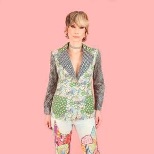 May include: A patterned blazer with a floral design in green, white, and blue. The sleeves have a small blue and white pattern. The blazer is paired with light-wash jeans with colorful patches. The background is a solid pink.