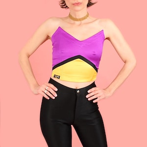May include: A colour block strapless top. The top has a purple upper section, a yellow mid-section, and a black accent. The model is wearing black trousers. The top has a small label on the front.