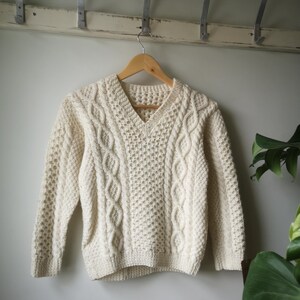 May include: A cream-coloured cable knit jumper with a v-neck. The jumper is hanging on a wooden hanger against a white wall.
