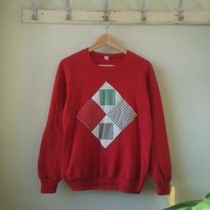 May include: Red sweatshirt with a diamond-shaped patchwork design on the front. The patchwork is made of different coloured fabrics, including red, green, white, and black.