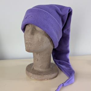 May include: A lavender fleece nightcap with a long, pointed tail. The hat is displayed on a mannequin head. The hat has a small embroidered design on the front. The hat is made of a soft, fuzzy material.