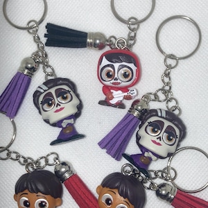 Könnte beinhalten: Schlüsselanhänger mit Figuren aus dem Animationsfilm Coco. Jeder Schlüsselanhänger hat einen silbernen Ring, eine Charakterfigur und eine Quaste. Die Figuren zeigen Charaktere in roten, lila und schwarzen Outfits.