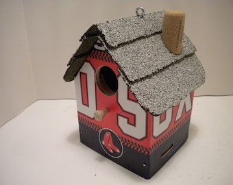 May include: A red, white, and blue birdhouse with a cork chimney and a baseball team logo. The birdhouse has a small opening for birds to enter and a perch for them to land on.