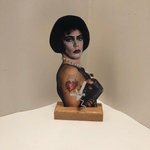 May include: A clock featuring a bust of a person with black hair and makeup, wearing a black top and a pearl necklace. The person has a tattoo of a broken heart with a dagger on their arm. The clock hands are white and the base is made of wood.