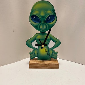 May include: A green alien figurine with large blue eyes and a clock face on its chest. The figurine is standing on a wooden base.