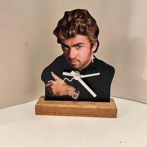 May include: A wooden clock featuring a black and white image of a man with brown hair. The clock has white hands and a black background. The clock is sitting on a wooden base.