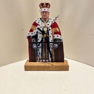 May include: A novelty clock featuring a figure in royal attire, complete with a crown and ornate robes. The clock hands are black, and the base is made of wood. The figure is standing on a balcony with a decorative railing.