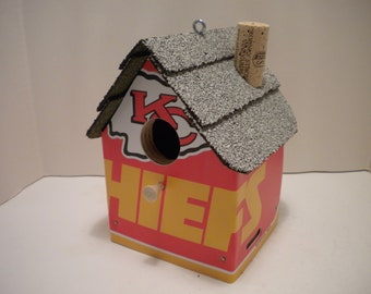 May include: A red and yellow birdhouse shaped like a house with a grey shingled roof. The front of the house has the letters "KC" and the word "CHIEFS" in yellow letters. A cork is on top of the roof.