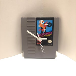 May include: A grey Nintendo Entertainment System game cartridge with a white clock face and hands. The game cartridge is for the game "Excitebike".