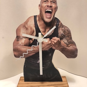 Puede incluir: Un reloj de madera con una imagen impresa de un hombre con un f&iacute;sico musculoso, vistiendo un tanque negro y una expresi&oacute;n decidida. Las manecillas del reloj son blancas y la base es de madera natural.