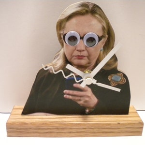 May include: A wooden clock with a picture of a woman wearing googly eye glasses. The clock has white hands and a wooden base.