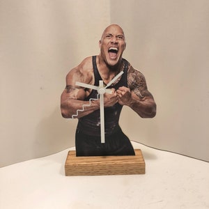 Puede incluir: Un reloj de madera con una imagen recortada de Dwayne "The Rock" Johnson, con una esfera y agujas blancas. El reloj est&aacute; sobre una base de madera.