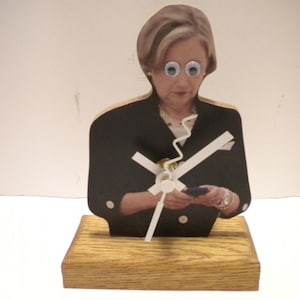 May include: A wooden clock featuring a printed image of a woman with googly eyes. The clock hands are white and the base is made of wood.