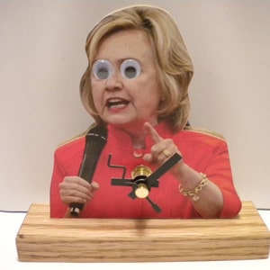 May include: A wooden clock with a red, white, and blue design. The clock features a picture of a woman with googly eyes and a microphone. The clock is on a wooden base.