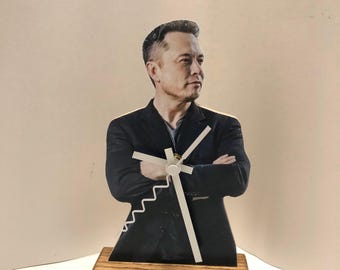 Puede incluir: Un reloj de madera con una imagen recortada de Elon Musk con una esfera y agujas blancas.