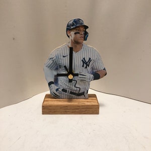 May include: A wooden clock featuring a baseball player in a blue and white striped uniform with a black and white baseball cap. The clock has black hands and a gold center.