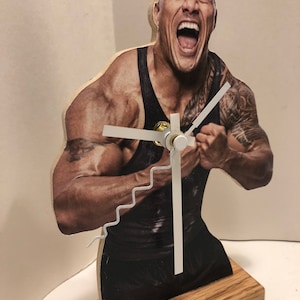 Puede incluir: Un reloj de madera con una foto de un hombre musculoso que lleva un tanque negro y una expresi&oacute;n decidida. Las manecillas del reloj son blancas y el reloj est&aacute; montado sobre una base de madera.