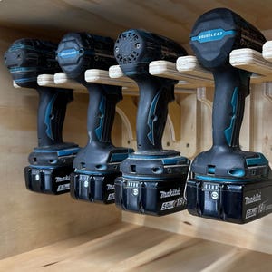 以下が含まれることがあります： 木製のラックに設置された、黒とティール色のコードレスドリル4台。バッテリーが取り付けられています。ドリルには「Brushless」と「Makita 5.0 18V」または「Makita 6.0 18V」のラベルが付いています。ラックは明るい色の木材でできています。