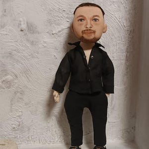 May include: A handmade doll with a painted face, wearing a black button-up shirt and black pants. The doll is standing on a white surface.