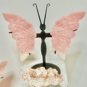 May include: A pink butterfly carved from stone is displayed on a black metal stand. The butterfly is sitting on a bed of white and pink crystals.
