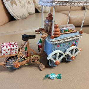 May include: A miniature wooden ice cream cart on a bicycle with a red and white striped gift box on the back. The cart has a white canopy with a lace trim and a sign that says "GELATO".