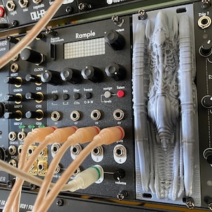 Può includere: Primo piano di un modulo sintetizzatore nero con manopole, pulsanti e jack. Il modulo è etichettato "Rample". Un modello 3D stampato di una creatura aliena è montato a destra del modulo.