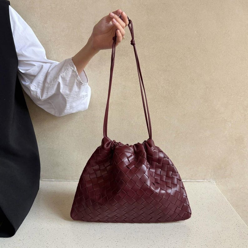 K&ouml;nnte beinhalten: Eine tiefburgunderrote Handtasche mit geflochtenem Lederau&szlig;enmaterial und einem gerafften Kordelzugverschluss. Die Tasche verf&uuml;gt &uuml;ber zwei d&uuml;nne, passende Schulterriemen aus Leder, die oben verknotet sind und eine weiche, lockere Form bilden.