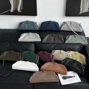 Op de afbeelding: Een verzameling geweven leren clutch tassen in verschillende kleuren, waaronder blauw, zilver, groen en bruin. De tassen zijn gerangschikt op een zwart oppervlak, waarbij hun verschillende tinten en texturen worden getoond. Elke tas heeft een sluiting en een schouderriem.