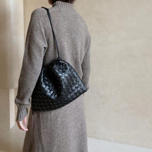 K&ouml;nnte beinhalten: Eine schwarze Umh&auml;ngetasche aus geflochtenem Leder mit einer weichen, lockeren Silhouette. Die Tasche verf&uuml;gt &uuml;ber einen d&uuml;nnen, integrierten Schulterriemen und eine geraffte &Ouml;ffnung an der Oberseite, die ein detailliertes Gittermuster zeigt.