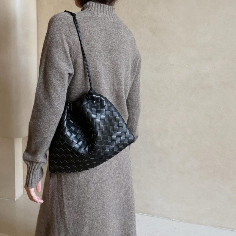 K&ouml;nnte beinhalten: Eine schwarze Umh&auml;ngetasche aus geflochtenem Leder mit einer weichen, lockeren Silhouette. Die Tasche verf&uuml;gt &uuml;ber einen d&uuml;nnen, integrierten Schulterriemen und eine geraffte &Ouml;ffnung an der Oberseite, die ein detailliertes Gittermuster zeigt.