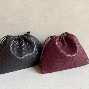 K&ouml;nnte beinhalten: Zwei weiche, beutelf&ouml;rmige Taschen mit geflochtenem Leder-Au&szlig;enmaterial. Eine Tasche ist dunkelbraun, die andere tiefburgunderrot. Beide Taschen haben einen Kordelzug am oberen Rand, der eine geraffte, abgerundete Form mit strukturierter Oberfl&auml;che erzeugt.