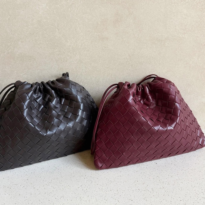 K&ouml;nnte beinhalten: Zwei weiche, beutelf&ouml;rmige Taschen mit geflochtenem Leder-Au&szlig;enmaterial. Eine Tasche ist dunkelbraun, die andere tiefburgunderrot. Beide Taschen haben einen Kordelzug am oberen Rand, der eine geraffte, abgerundete Form mit strukturierter Oberfl&auml;che erzeugt.