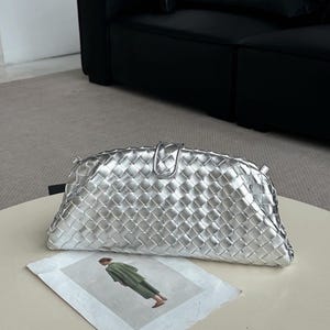 Op de afbeelding: Een zilveren clutch met een geweven ontwerp en een klepsluiting. De tas ligt op een lichtgekleurde tafel, met een gedrukte afbeelding van een persoon in groene kleding.