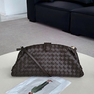 Op de afbeelding: Een donkerbruine geweven leren clutch met een schouderriem. De tas heeft een rechthoekige vorm met een gebogen bovenkant en een klepsluiting. De tas ligt op een witte tafel.