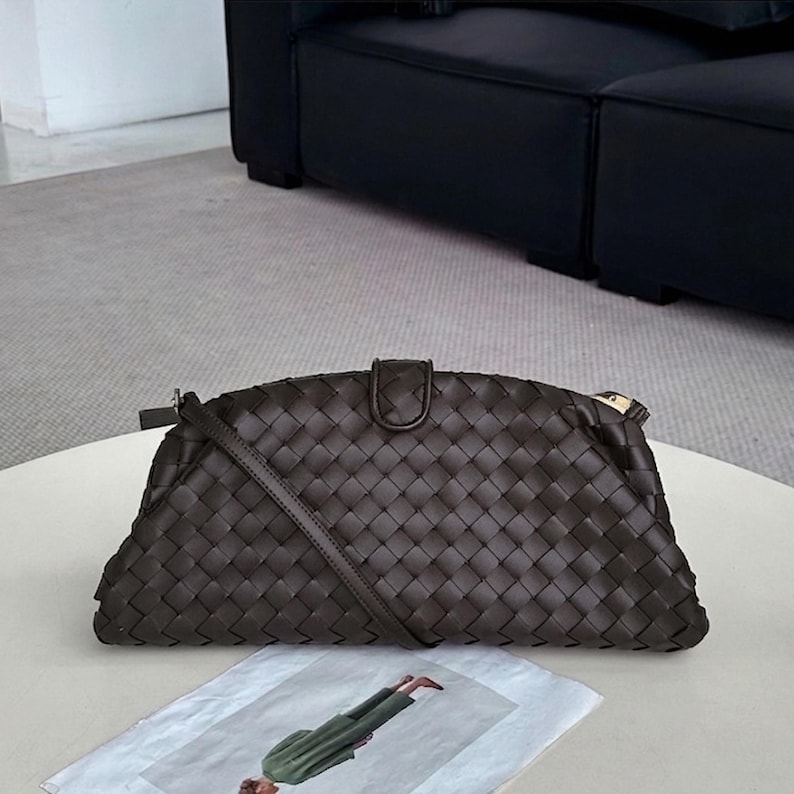 Op de afbeelding: Een donkerbruine geweven leren clutch met een schouderriem. De tas heeft een rechthoekige vorm met een gebogen bovenkant en een klepsluiting. De tas ligt op een witte tafel.
