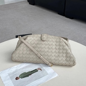 Op de afbeelding: Een lichtbeige geweven clutch tas met een lange, dunne riem en een goudkleurige gesp. De tas heeft een afgeronde vorm en een klepsluiting. Een afgedrukte afbeelding van een persoon in groene kleding ligt op de tafel.