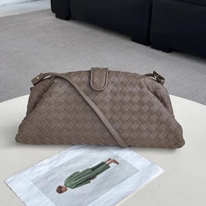 Op de afbeelding: Een taupekleurige geweven leren handtas met een schouderriem. De tas heeft een rechthoekige vorm met een gebogen bovenkant en een klepsluiting. De tas ligt op een wit oppervlak met een mode-illustratie.