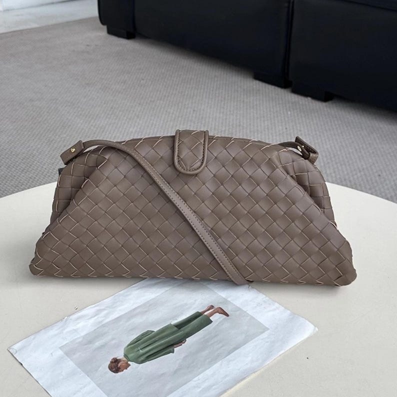 Op de afbeelding: Een taupekleurige geweven leren handtas met een schouderriem. De tas heeft een rechthoekige vorm met een gebogen bovenkant en een klepsluiting. De tas ligt op een wit oppervlak met een mode-illustratie.