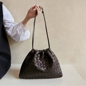 K&ouml;nnte beinhalten: Eine dunkelbraune Ledertasche mit einem durchgehenden geflochtenen Korbgeflechtmuster. Die Tasche verf&uuml;gt &uuml;ber einen gerafften Verschluss im Kordelzugstil und zwei d&uuml;nne, lange Schulterriemen, die oben f&uuml;r einen sicheren Halt verknotet sind.