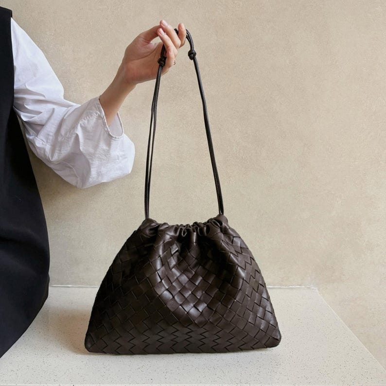 K&ouml;nnte beinhalten: Eine dunkelbraune Ledertasche mit einem durchgehenden geflochtenen Korbgeflechtmuster. Die Tasche verf&uuml;gt &uuml;ber einen gerafften Verschluss im Kordelzugstil und zwei d&uuml;nne, lange Schulterriemen, die oben f&uuml;r einen sicheren Halt verknotet sind.