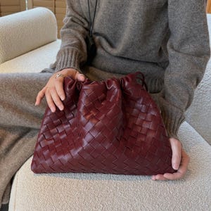 K&ouml;nnte beinhalten: Eine tiefburgunderrote Beuteltasche mit geflochtenem Lederau&szlig;enmaterial und einem gerafften Kordelzugverschluss an der Oberseite. Die Tasche hat eine weiche, flexible Struktur und ein gleichm&auml;&szlig;iges Gittermuster aus ineinandergreifenden Lederstreifen.