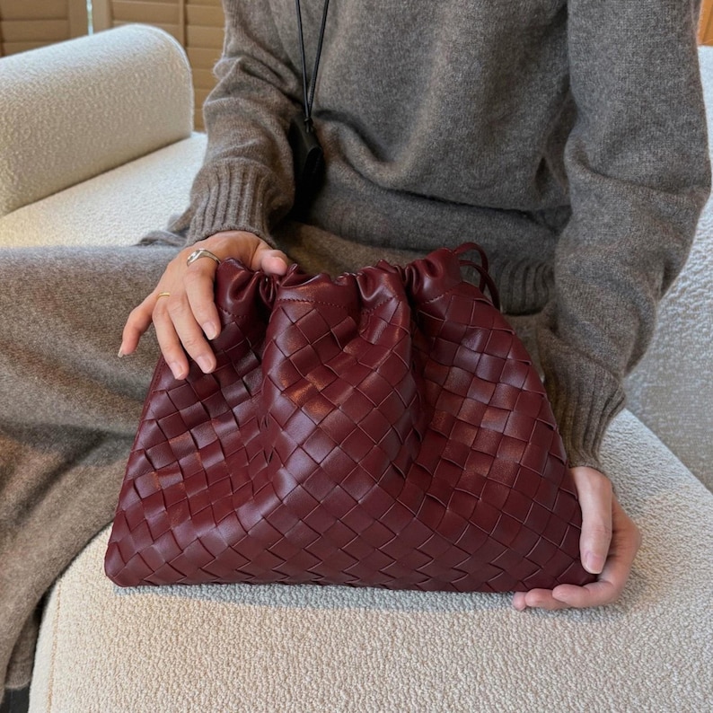 K&ouml;nnte beinhalten: Eine tiefburgunderrote Beuteltasche mit geflochtenem Lederau&szlig;enmaterial und einem gerafften Kordelzugverschluss an der Oberseite. Die Tasche hat eine weiche, flexible Struktur und ein gleichm&auml;&szlig;iges Gittermuster aus ineinandergreifenden Lederstreifen.