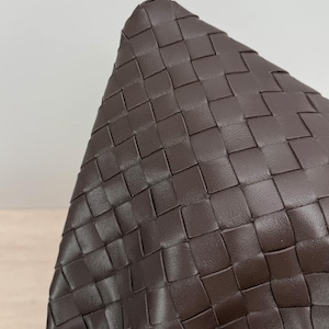 K&ouml;nnte beinhalten: Eine dunkelbraune Ledertasche mit einem geflochtenen Intrecciato-Muster. Die Oberfl&auml;che besteht aus gleichm&auml;&szlig;igen, ineinandergreifenden Streifen aus glattem Leder, die eine konsistente gitterartige Textur auf der gesamten sichtbaren Au&szlig;enseite bilden.