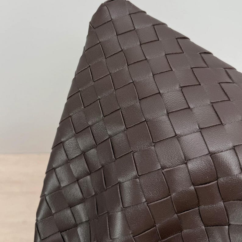 K&ouml;nnte beinhalten: Eine dunkelbraune Ledertasche mit einem geflochtenen Intrecciato-Muster. Die Oberfl&auml;che besteht aus gleichm&auml;&szlig;igen, ineinandergreifenden Streifen aus glattem Leder, die eine konsistente gitterartige Textur auf der gesamten sichtbaren Au&szlig;enseite bilden.