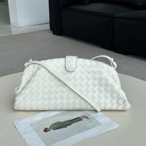 Op de afbeelding: Een witte, geweven leren schoudertas met een klepsluiting en een lange riem. De tas is rechthoekig met afgeronde randen. Een tijdschrift met een persoon in groene kleding staat op de voorgrond.