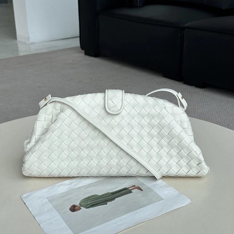 Op de afbeelding: Een witte, geweven leren schoudertas met een klepsluiting en een lange riem. De tas is rechthoekig met afgeronde randen. Een tijdschrift met een persoon in groene kleding staat op de voorgrond.